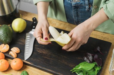 Muz soyan, ıspanak yapraklı, elmalı, mandalinalı ve avokadolu sağlıklı vejetaryen kokteyli hazırlayan bir kadın..
