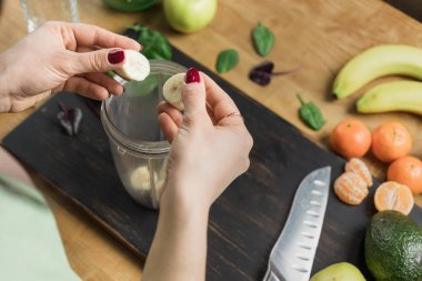 Elinde muz parçaları olan bir kadın, masada ıspanaklı, elmalı ve avokadoyla sağlıklı vejetaryen kokteyli hazırlıyor..