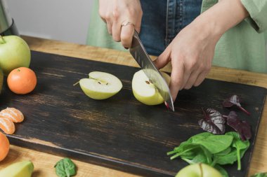 Elma kesen kadın, masada ıspanak yapraklı, muzlu ve avokadolu sağlıklı vejetaryen smoothie hazırlıyor..