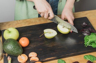 Elma kesen kadın, masada ıspanak yapraklı, muzlu ve avokadolu sağlıklı vejetaryen smoothie hazırlıyor..