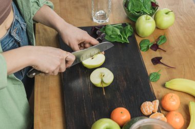 Elma kesen kadın, masada ıspanak yapraklı, muzlu ve avokadolu sağlıklı vejetaryen smoothie hazırlıyor..