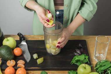 Elma parçaları tutan bir kadın, masada ıspanaklı, muzlu ve avokadolu sağlıklı vejetaryen smoothie hazırlıyor..