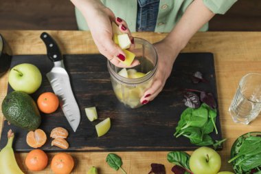 Elma parçaları tutan bir kadın, masada ıspanaklı, muzlu ve avokadolu sağlıklı vejetaryen smoothie hazırlıyor..