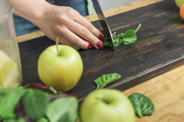 Yeşil ıspanak yaprakları kesen bir kadın, sağlıklı vejetaryen smoothie hazırlarken ıspanak, muz, elma ve avokado karıştırıcıyı masaya koyar..