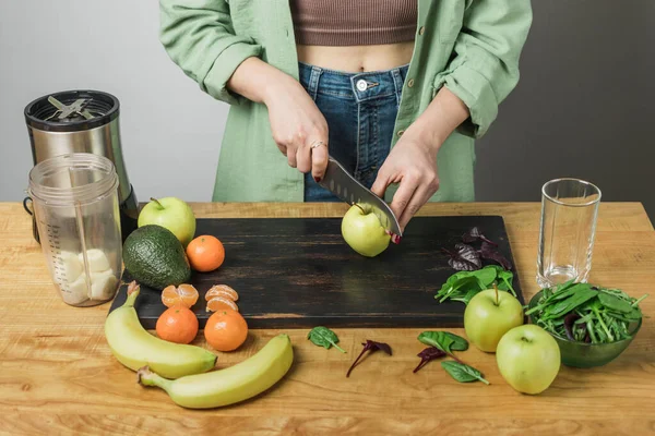 Elma kesen kadın, masada ıspanak yapraklı, muzlu ve avokadolu sağlıklı vejetaryen smoothie hazırlıyor..