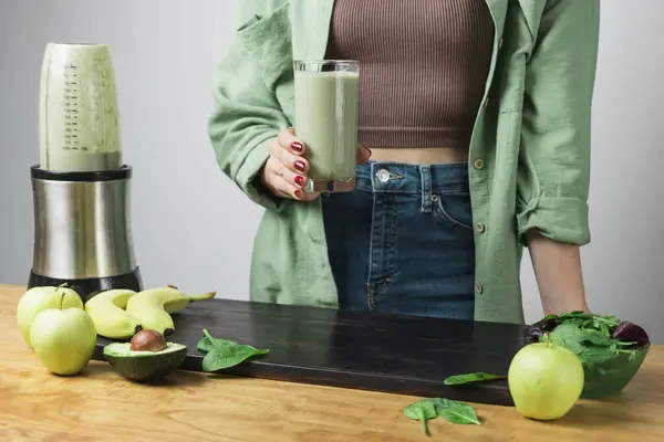 Tanımlanamayan sıradan bir kadın elinde bir bardak taze, sağlıklı, yeşil vejetaryen meyve püresi tutuyor. muz, ıspanak, elma ve avokado bir masada..