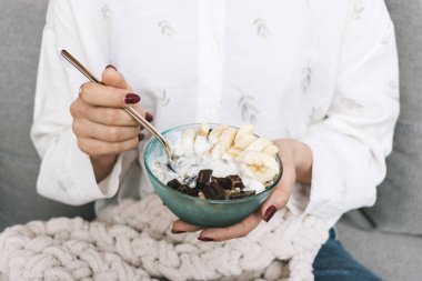 Kanepede oturan ve kahvaltıda sağlıklı yiyecekler yiyen bir kadın, mısır gevreği, granola, müsli, muzlu yulaf ezmesi, siyah çikolata ve yoğurt..