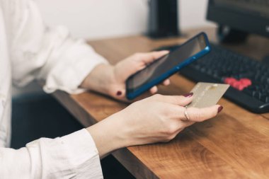 Kadının elleri kredi kartını tutuyor ve online alışveriş, ödeme veya çalışma için cep telefonu ve bilgisayar kullanıyor.