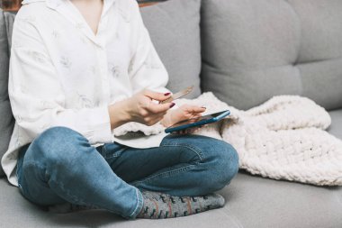 Kanepede oturan tanınmayan bir kadın, elinde kredi kartı, cep telefonu, internet alışverişi ya da internet ödemesi için akıllı telefon kullanıyor..