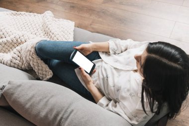 Güzel bir genç kadın kanepede uzanıyor ve beyaz ekranlı cep telefonu kullanıyor. Sosyal medya için akıllı telefon, internet taraması veya mesajlaşma..