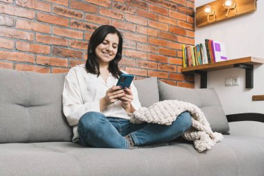 Kanepede oturmuş gülümseyen güzel bir genç kadın cep telefonu kullanıyor, sosyal medya için akıllı telefon, internette geziniyor ya da mesajlaşıyor..