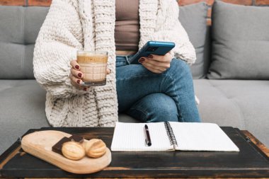 Evdeki koltukta oturan tanınmayan bir kadın cep telefonu ya da akıllı telefon kullanıyor ve elinde bir bardak kahve tutuyor. Tatlı ve masanın üzerinde bir kalem olan not defteri..