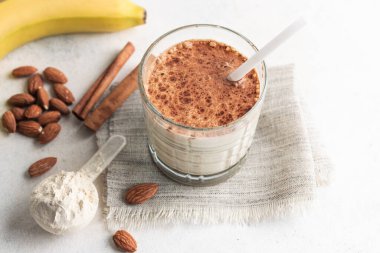 Proteinli içecek, muzlu beyaz masada sağlıklı milkshake, ölçü kaşığında protein tozu, badem fıstığı ve tarçın çubukları..