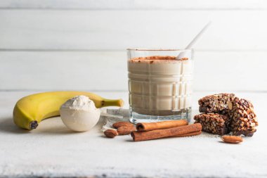 Protein içeceği, muzlu beyaz masada sağlıklı milkshake, ölçü kaşığında protein tozu, protein barı, badem fıstığı ve tarçın çubukları..