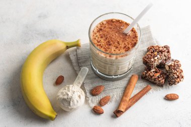 Protein içeceği, muzlu beyaz masada sağlıklı milkshake, ölçü kaşığında protein tozu, protein barı, badem fıstığı ve tarçın çubukları..