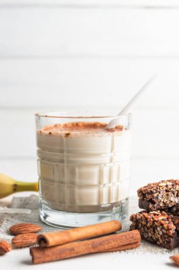 Protein içeceği, muzlu beyaz masada sağlıklı milkshake, ölçü kaşığında protein tozu, protein barı, badem fıstığı ve tarçın çubukları..