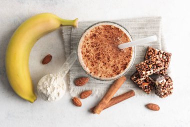 Protein içeceği, muzlu beyaz masada sağlıklı milkshake, ölçü kaşığında protein tozu, protein barı, badem fıstığı ve tarçın çubukları..