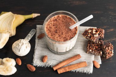 Protein içeceği, ahşap tahtada sağlıklı milkshake smoothie muz, protein tozu ölçme kaşığı, protein barı, badem fıstığı ve tarçın çubukları..