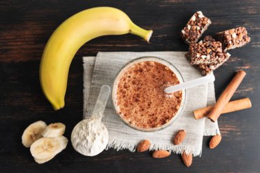 Protein içeceği, ahşap tahtada sağlıklı milkshake smoothie muz, protein tozu ölçme kaşığı, protein çubuğu, badem fıstığı ve tarçın çubukları, üst görüş..