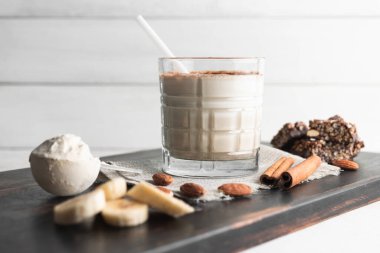 Protein içeceği, ahşap tahtada sağlıklı milkshake smoothie muz, protein tozu ölçme kaşığı, protein barı, badem fıstığı ve tarçın çubukları..
