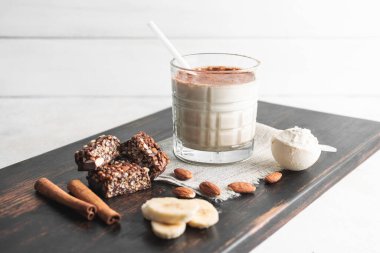 Protein içeceği, ahşap tahtada sağlıklı milkshake smoothie muz, protein tozu ölçme kaşığı, protein barı, badem fıstığı ve tarçın çubukları..