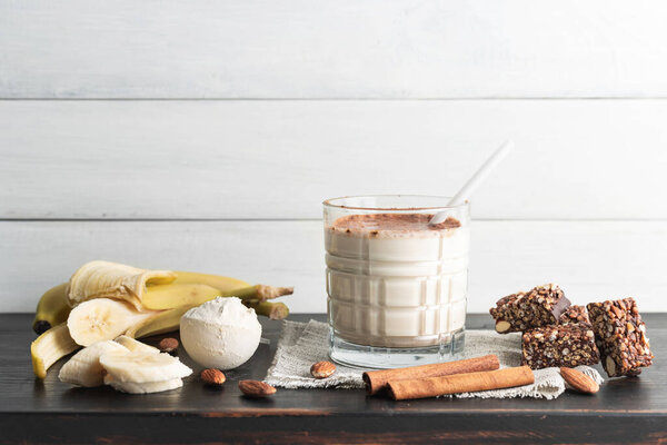 Glass with protein drink, healthy milkshake smoothie on wooden board with bananas, protein powder in measuring spoon, protein bar, almond nuts and cinnamon sticks.