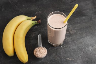 Sarı pipetli bir bardakta çikolatalı protein içeceği. Siyah arka planda protein tozu ve muzla plastik ölçüm kaşığı..