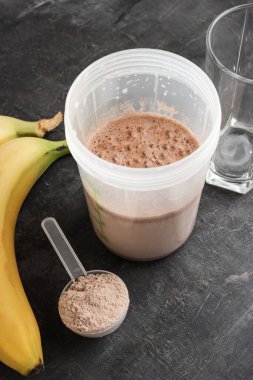 Karışık çikolatalı protein içeceği, plastik ölçüm kaşığı protein tozu, muz ve koyu bir arka planda içki bardağı..