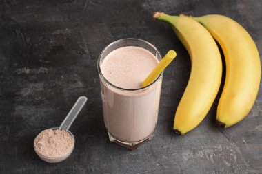 Sarı pipetli bir bardakta çikolatalı protein içeceği. Siyah arka planda protein tozu ve muzla plastik ölçüm kaşığı..