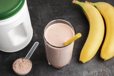 Sarı pipetli bir bardakta çikolatalı protein içeceği. Siyah arka planda protein tozu ve muzla plastik ölçüm kaşığı..