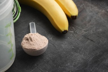Plastik bir kaşıkta çikolata aromalı beyaz protein tozu. Arka planda, muzlu ve sallanan bir kaşık..