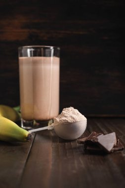 Sütlü protein tozlu plastik kaşık, bardakta çikolatalı milkshake kokteyli, harmanlanmış protein içeceği ve ahşap arka planda muz meyvesi..