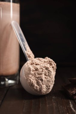 Protein tozlu plastik ölçü kaşığı, bardakta çikolatalı milkshake kokteyli, koyu ahşap arka planda karışık protein içeceği..