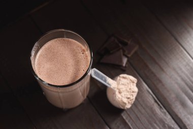 Sütlü protein tozlu plastik ölçüm kaşığı, bardakta milkshake kokteyli, karışık protein içeceği ve koyu ahşap arka planda çikolata küpleri..
