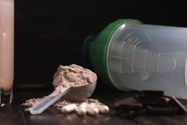 Protein tozlu plastik kaşık, bardakta milkshake kokteyli, karışık protein içeceği, beyaz haplar veya kapsüller, koyu ahşap arka planda çikolata küpleri..