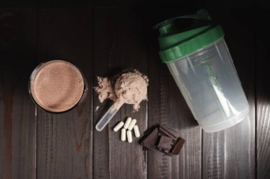Bardakta protein milkshake kokteyli, beyaz protein tozlu plastik ölçüm kaşığı, beyaz haplar veya amino asitlerin kapsülleri, koyu ahşap arka planda çikolata küpleri, üst görünüm..