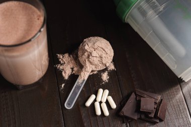 Protein tozlu plastik kaşık, bardakta milkshake kokteyli, karışık protein içeceği, beyaz haplar veya kapsüller, koyu ahşap arka planda çikolata küpleri..