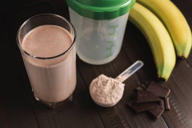 Bardakta protein milkshake kokteyli, beyaz protein tozuyla plastik ölçüm kaşığı, karıştırılmış protein içeceği, çikolata küpleri ve muz meyvesi hazırlamak için karıştırıcı koyu ahşap arka planda..