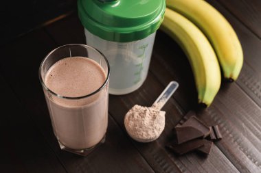 Bardakta protein milkshake kokteyli, beyaz protein tozuyla plastik ölçüm kaşığı, karıştırılmış protein içeceği, çikolata küpleri ve muz meyvesi hazırlamak için karıştırıcı koyu ahşap arka planda..