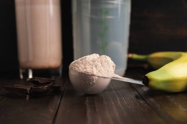 Sütlü protein tozlu plastik ölçüm kaşığı, bardakta milkshake kokteyli, karıştırılmış protein içeceği, çikolata küpleri ve muz meyvesi hazırlamak için karıştırıcı koyu ahşap arka planda.