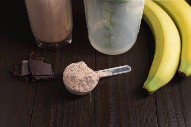 Sütlü protein tozlu plastik ölçüm kaşığı, bardakta milkshake kokteyli, karıştırılmış protein içeceği, çikolata küpleri ve muz meyvesi hazırlamak için karıştırıcı koyu ahşap arka planda.