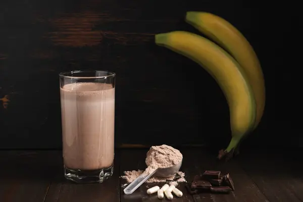 Plastic measuring spoon with whey protein powder, milkshake cocktail in a glass, blended protein drink, white pills or capsules, chocolate cubes and banana fruit on a dark wooden background.