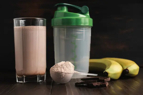 Plastic measuring spoon with whey protein powder, milkshake cocktail in a glass, shaker for prepare blended protein drink, chocolate cubes and banana fruit on a dark wooden background.