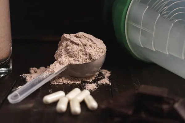 Plastic measuring spoon with whey protein powder, milkshake cocktail in a glass, blended protein drink, white pills or capsules, chocolate cubes on a dark wooden background.