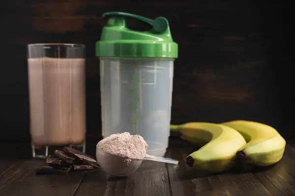 Plastic measuring spoon with whey protein powder, milkshake cocktail in a glass, shaker for prepare blended protein drink, chocolate cubes and banana fruit on a dark wooden background.