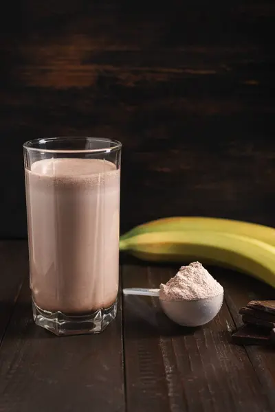 Plastic measuring spoon with whey protein powder, chocolate milkshake cocktail in a glass, blended protein drink and banana fruit on a dark wooden background.