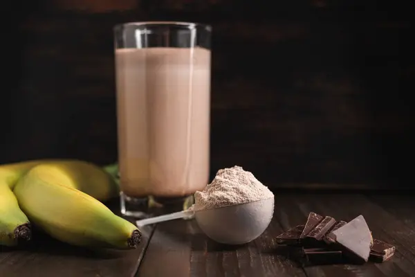 Plastic measuring spoon with whey protein powder, chocolate milkshake cocktail in a glass, blended protein drink and banana fruit on a dark wooden background.