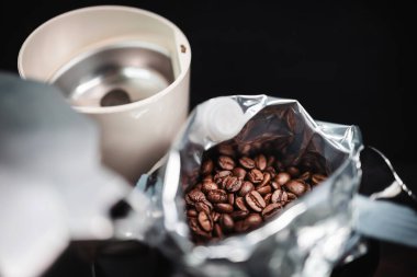 Kavrulmuş kahve çekirdekleri, açık bir folyo torbasında, üzerinde elektrikli kahve öğütücüsü ve bir moka demliği olan bir masada..