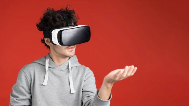 Genç bir adam VR gözlüklerini kullanarak oyun oynuyor, elinde sanal bir şey tutuyor, kırmızı arka plan.