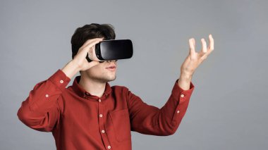 Genç bir adam sanal gerçeklik gözlüğü kullanarak oyun oynuyor, elinde gri bir arka plan var.